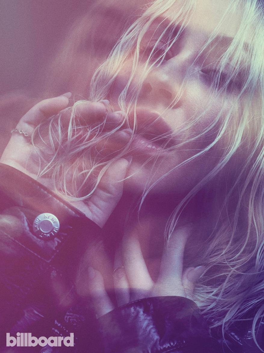 Woman with blond hair poses with her hands near her face. She is wearing a black jacket. Billboard logo is visible in corner.