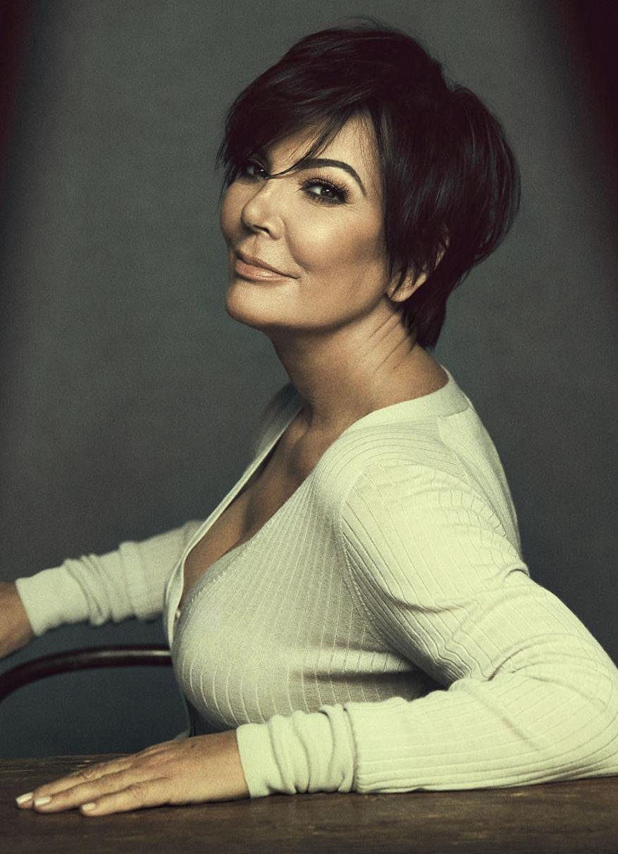 Celebrity portrait of Kris Jenner in long-sleeved top, seated at a wooden table in front of a grey backdrop, looking at the camera.