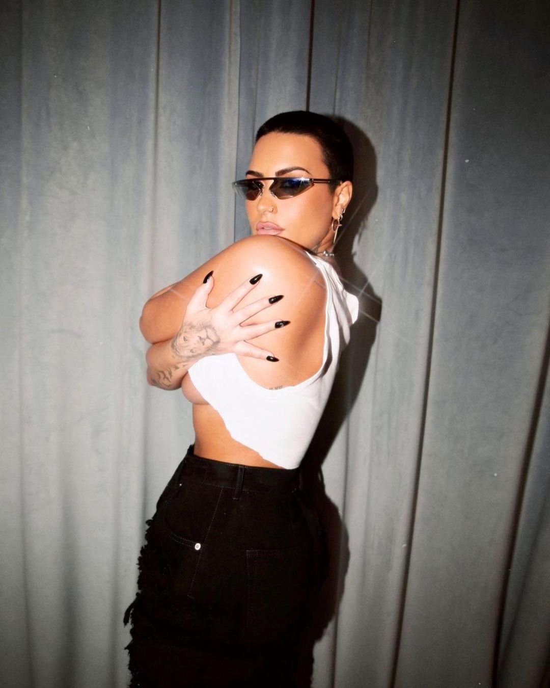 Woman with sunglasses poses in front of a grey curtain. She has tattoos on her arms and is wearing a white top and black pants.