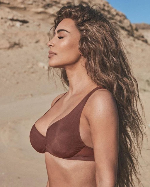 Side-view of an adult female with long brown hair wearing a brown bikini top against a sandy beach and blue sky background.