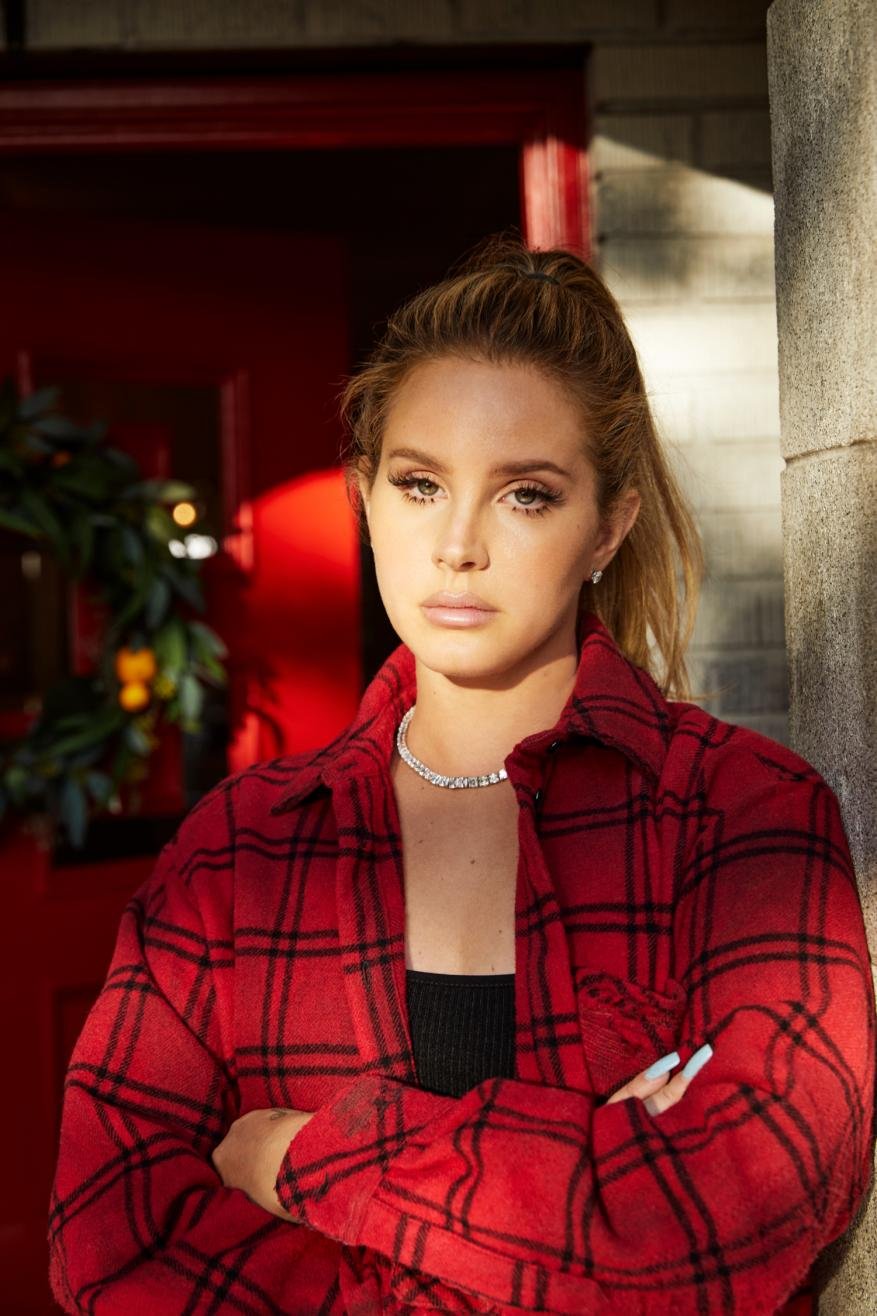 Portrait of a blonde woman in a red plaid flannel shirt with arms crossed in front of a red door decorated with greenery.