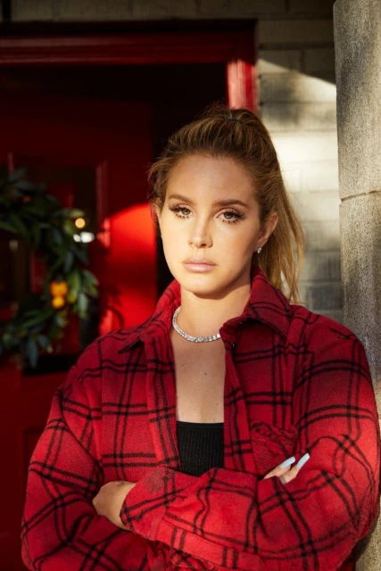 Portrait of a blonde woman in a red plaid flannel shirt with arms crossed in front of a red door decorated with greenery.