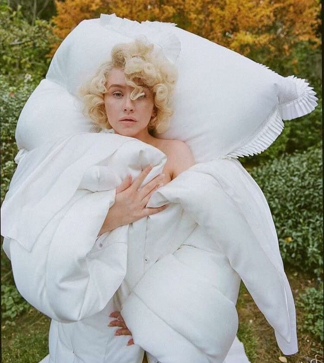 Woman with blonde curly hair, posing in a draped white textile against an outdoor backdrop with green foliage and autumn leaves.