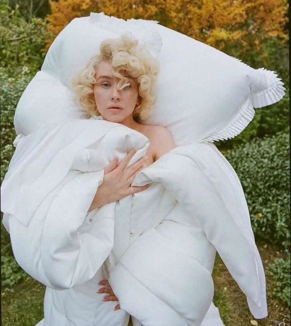 Woman with blonde curly hair, posing in a draped white textile against an outdoor backdrop with green foliage and autumn leaves.