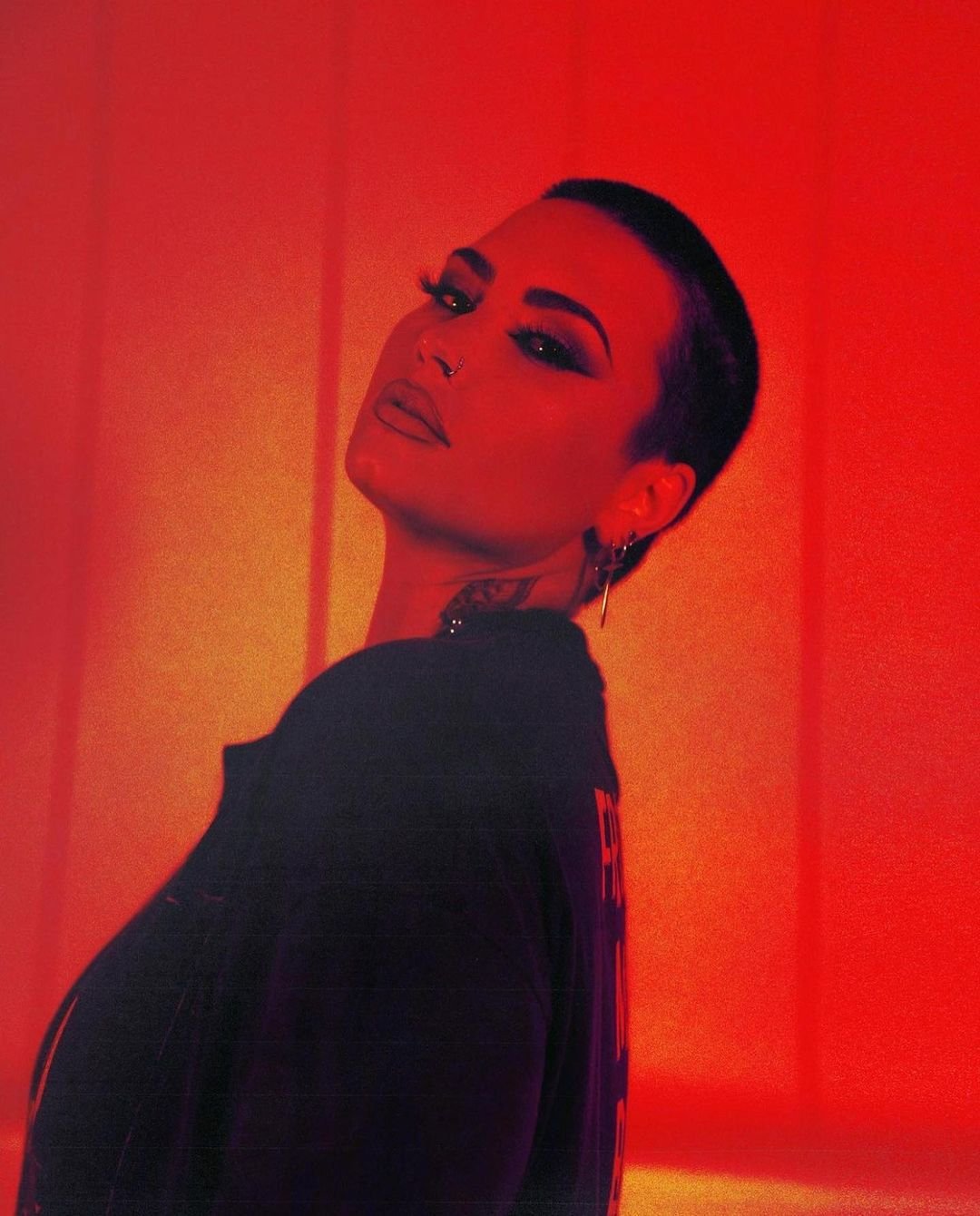 Close-up portrait of a person with short dark hair, heavy makeup, and visible neck tattoo, set against a striking red backdrop.