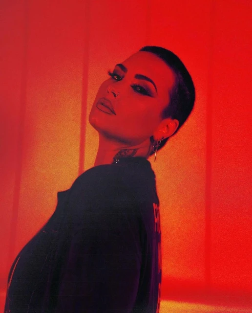 Close-up portrait of a person with short dark hair, heavy makeup, and visible neck tattoo, set against a striking red backdrop.