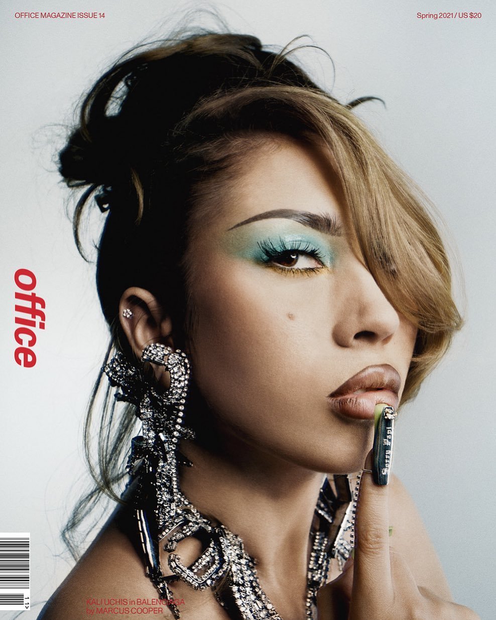 Close-up of a woman with glamorous makeup, including vibrant blue eyeshadow and glossy lips, posing for a fashion magazine cover with large statement earrings.