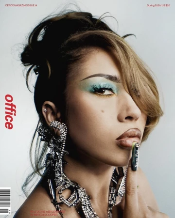 Close-up of a woman with glamorous makeup, including vibrant blue eyeshadow and glossy lips, posing for a fashion magazine cover with large statement earrings.