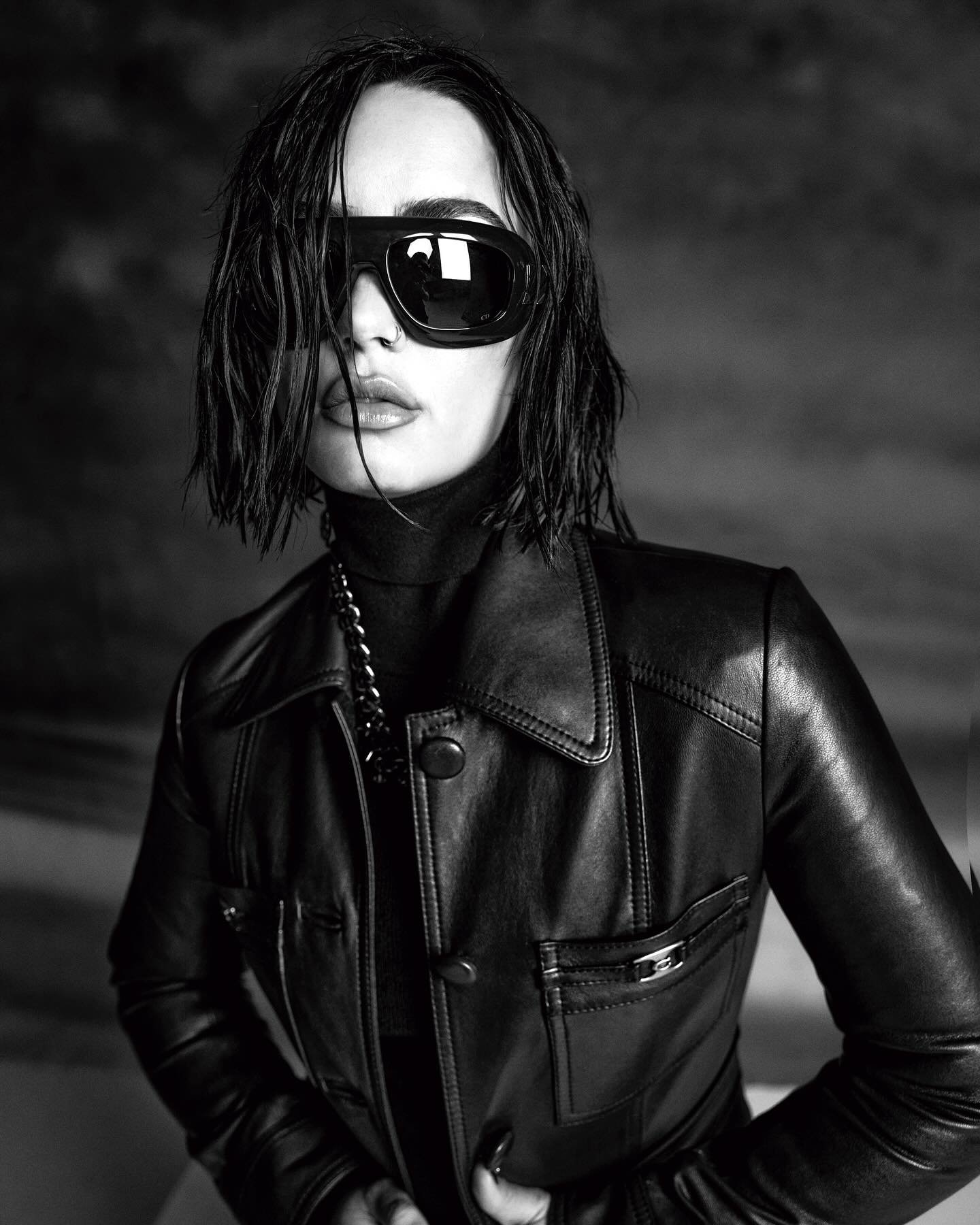 Black and white portrait of a woman with short hair wearing a black leather jacket and sunglasses against a blurred backdrop.