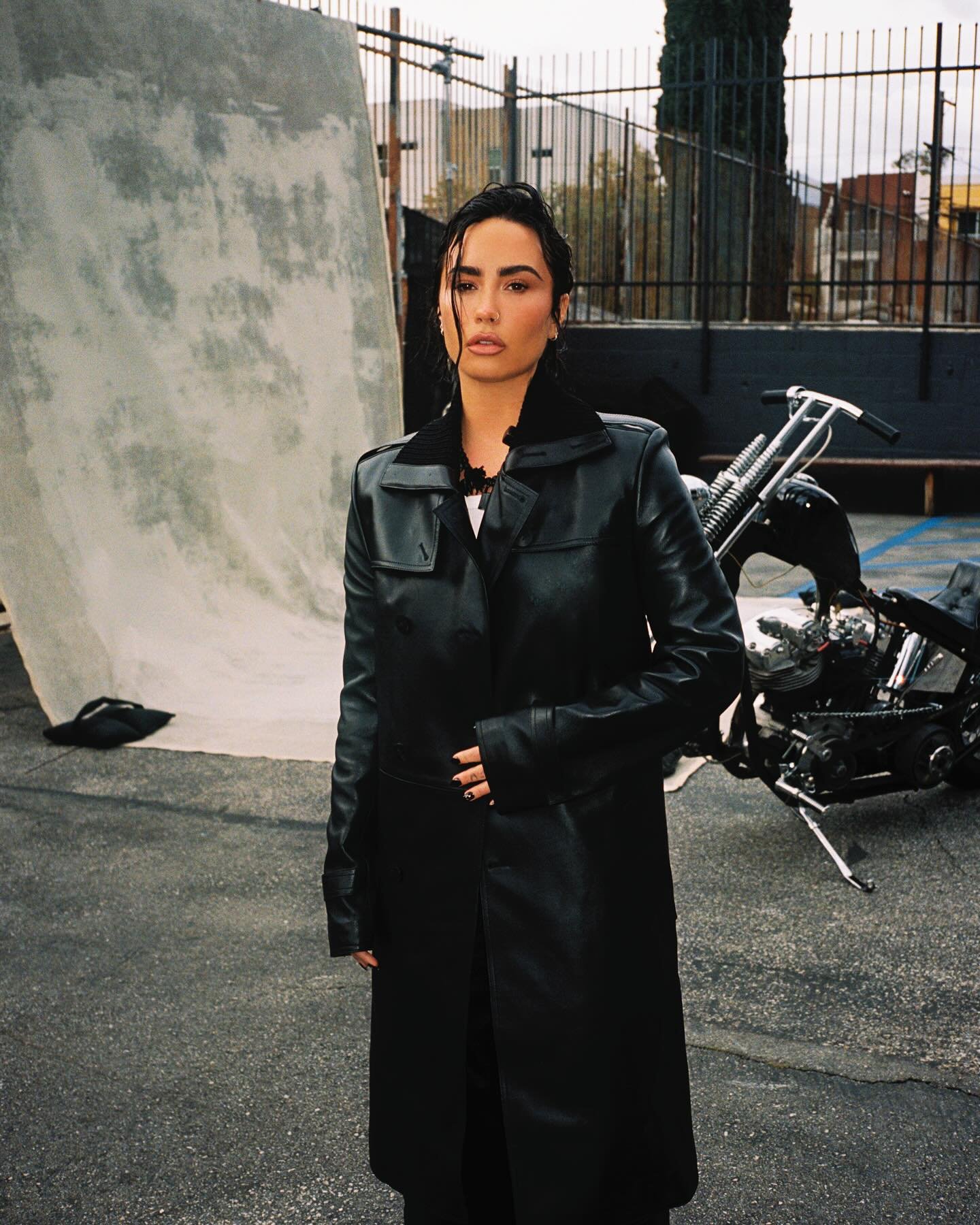 Demi Lovato poses in a black trench coat on a city street with a motorcycle and a backdrop. Moody outdoor fashion shot.