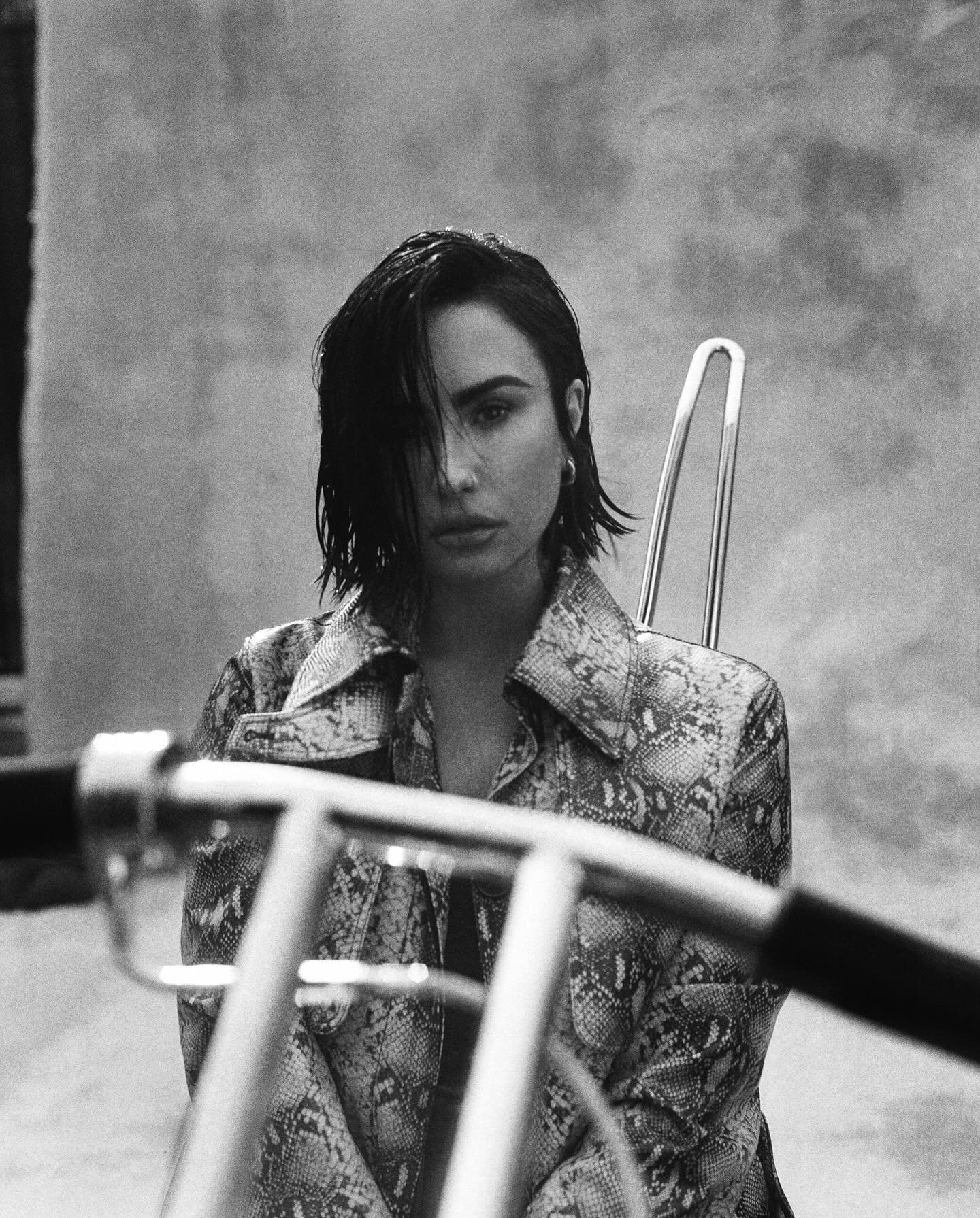 Black and white portrait of a woman in an animal-print jacket, standing in front of a textured gray wall with a bicycle in the foreground.