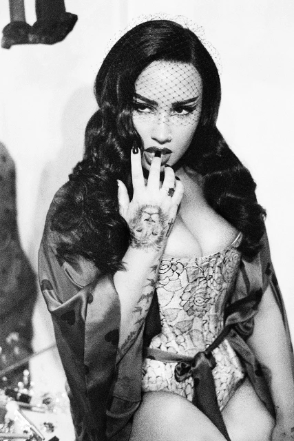 Black and white portrait of woman in floral corset and robe with finger to her lips and lion tattoo on her arm in an indoor setting.