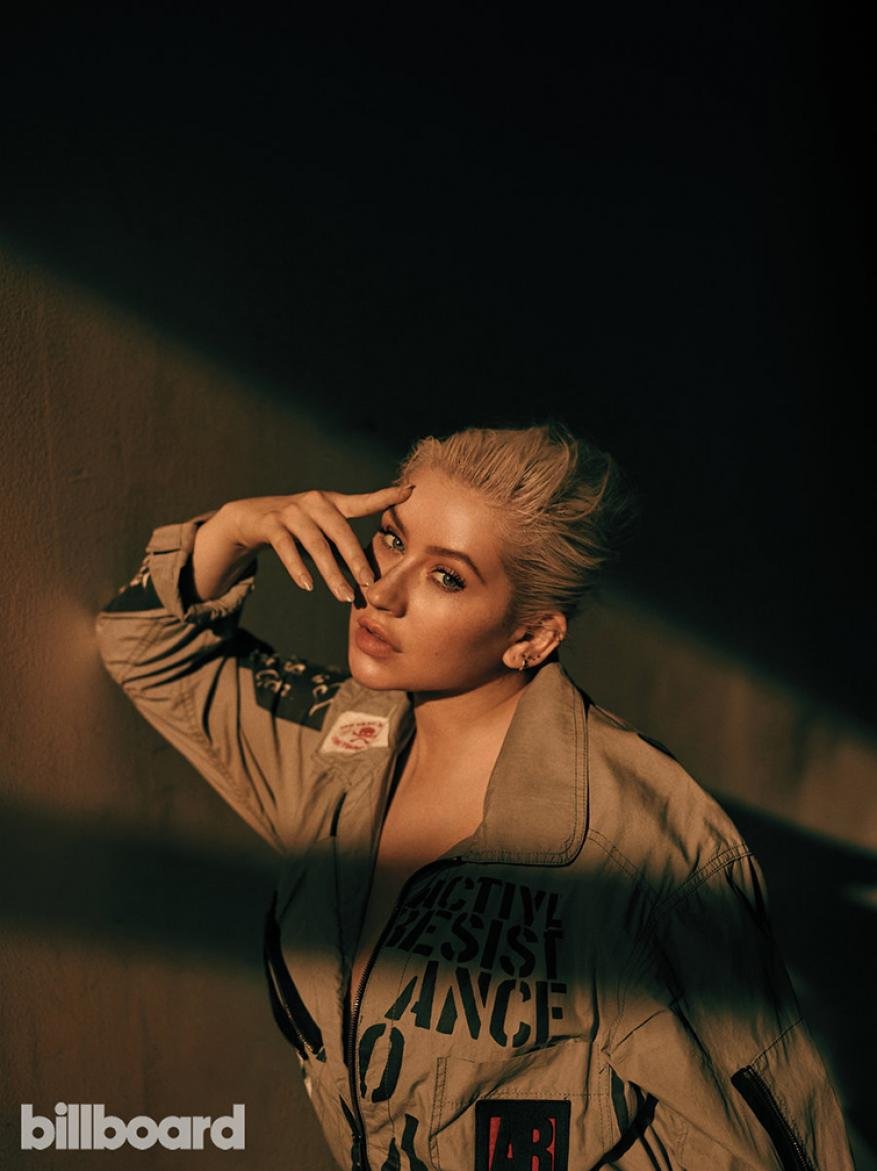 Christina Aguilera poses in a khaki flight suit with graphics, bathed in a warm spotlight against a dark background in a medium shot.