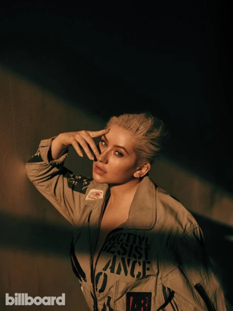 Christina Aguilera poses in a khaki flight suit with graphics, bathed in a warm spotlight against a dark background in a medium shot.