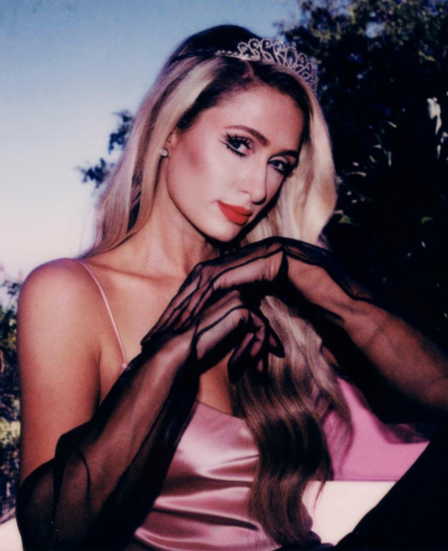 Portrait of an adult female with blond hair, wearing a tiara and sheer black gloves, posing in front of dark green foliage.