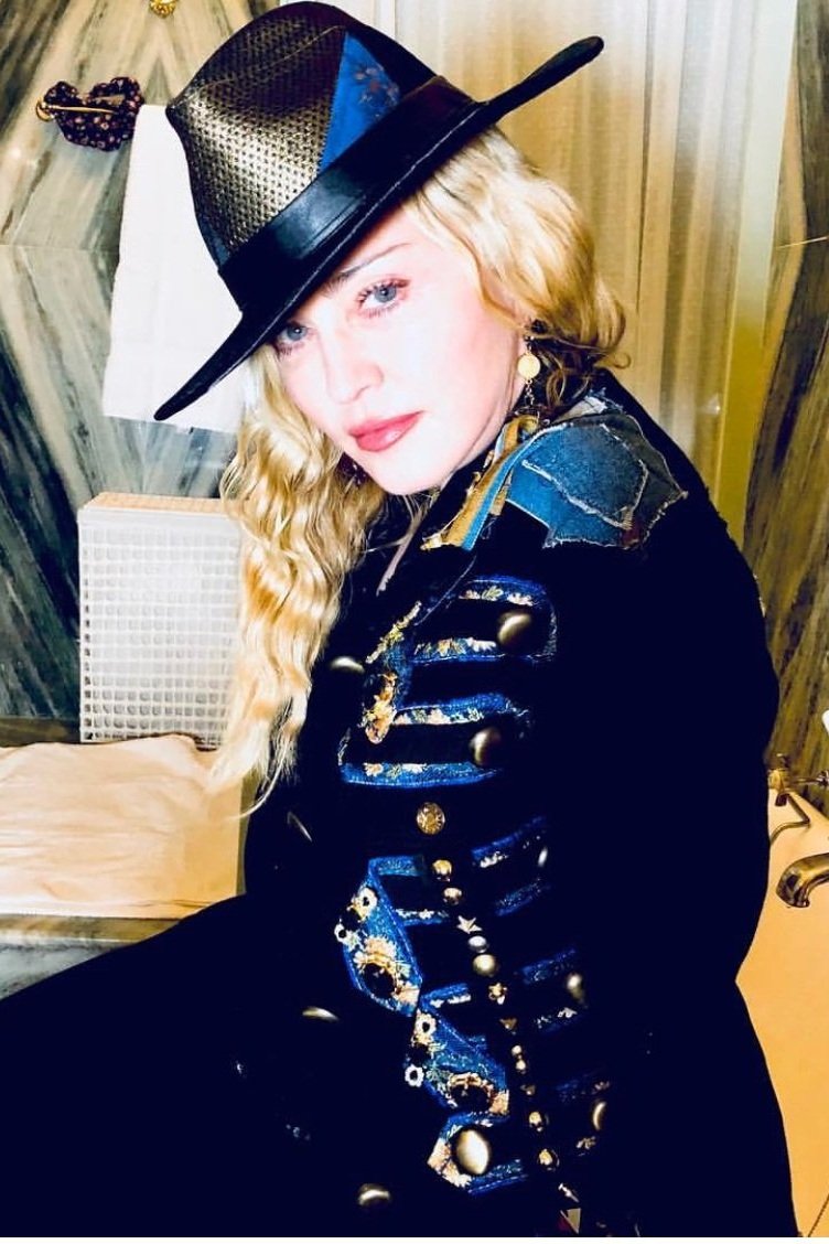 Portrait of Madonna wearing a black coat with blue accents, a custom hat with gold trim, in an indoor setting with a marble wall.
