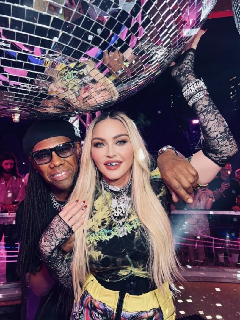 Madonna and a man pose beneath a disco ball in a selfie-style portrait, with stage lights and a crowd visible in the background.