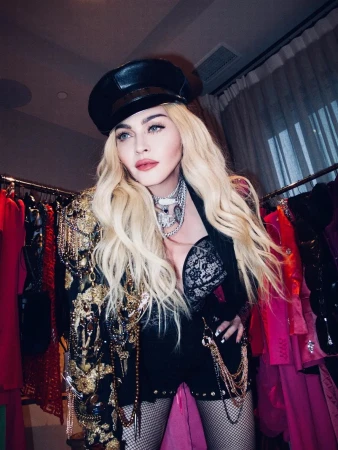 Close-up of a woman wearing a hat and black lace top with a black jacket and heavy gold jewelry posing indoors with racks of clothing.