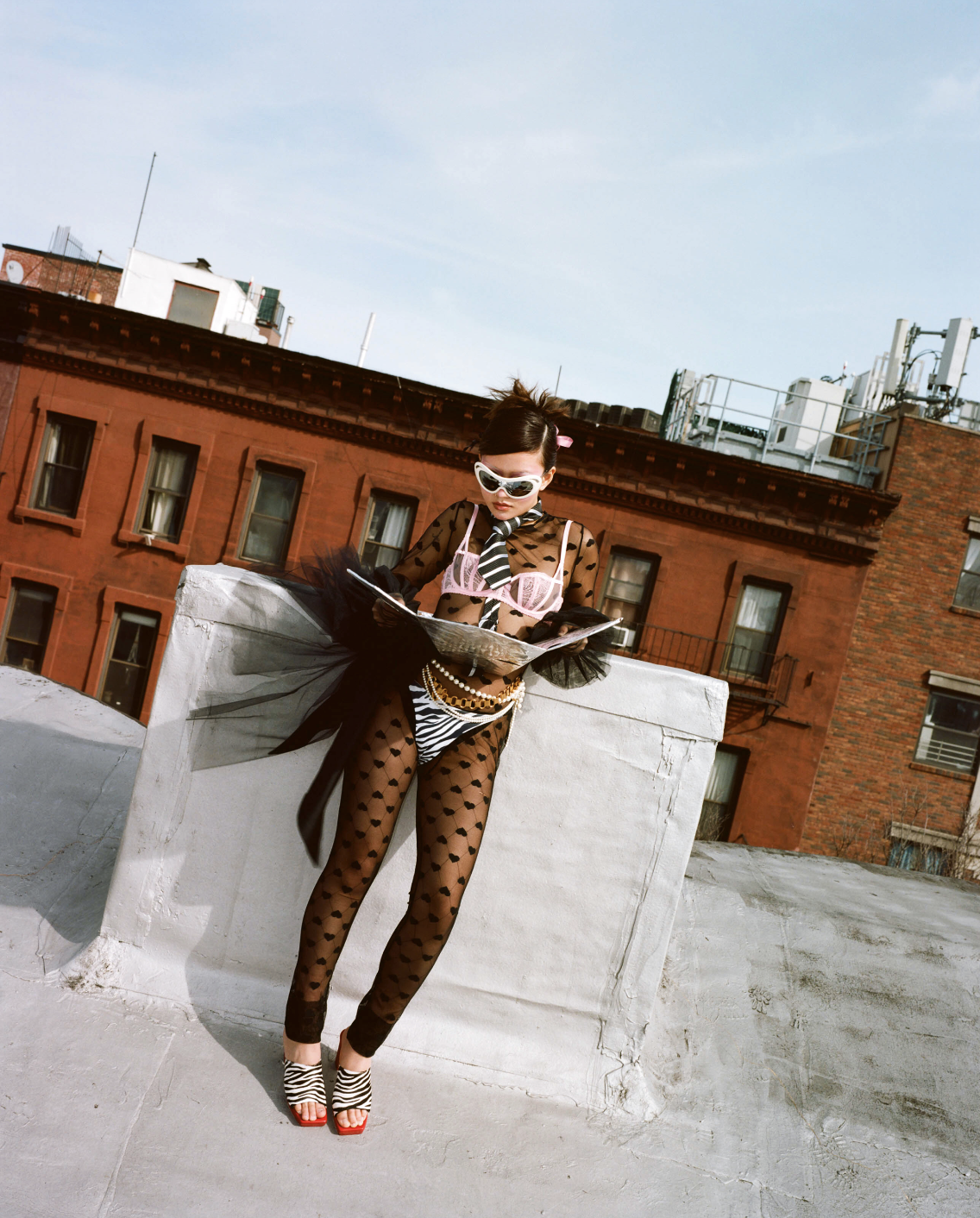 A fashionable woman in patterned clothing poses with a newspaper on a rooftop with red brick buildings in the background on a sunny day.