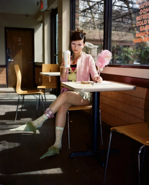 Young woman with creative makeup sits at a cafe table holding a drink and pen, wearing a pink cardigan and patterned top indoors.