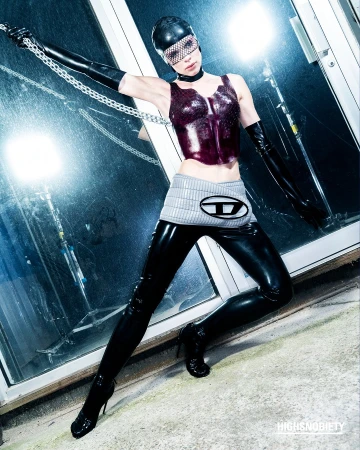 A woman in a latex outfit poses indoors with chains and a Diesel logo, showcasing a high-fashion, edgy style against a blurred, industrial backdrop.