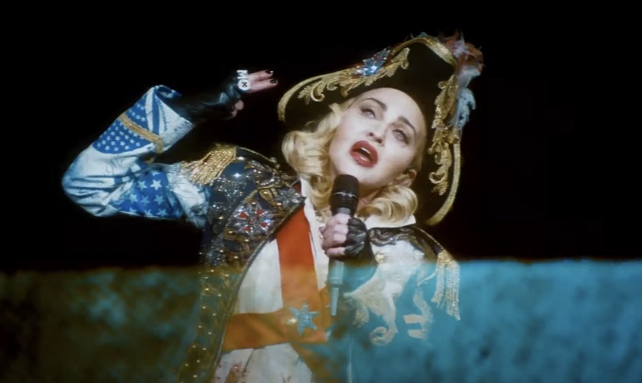 Madonna performs on stage wearing a pirate-themed outfit with a hat and microphone, set against a dark background.