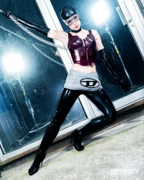 Model wearing a dark purple top, shiny black pants, and a mesh visor cap holding a metal chain in a studio setting