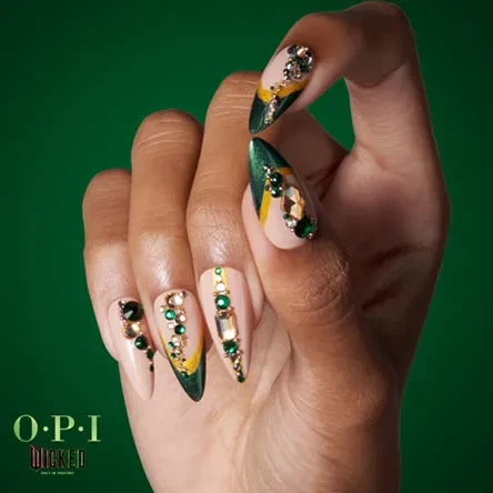 Close-up of a hand with long, almond-shaped nails decorated with rhinestones, green and gold polish, set against a green background.