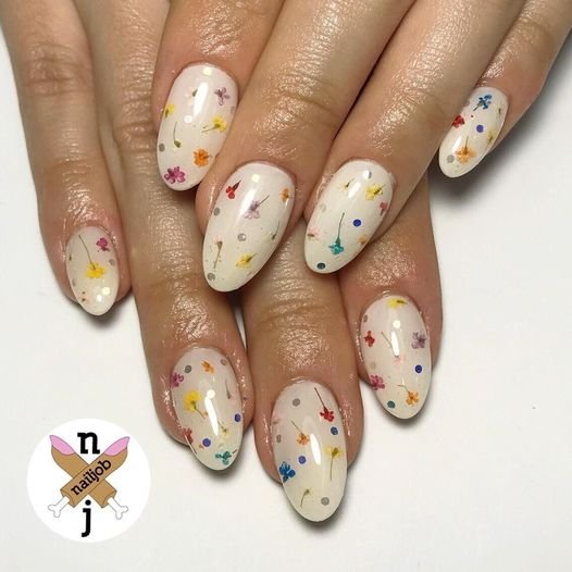 Close-up of manicured hand showcasing oval-shaped nails adorned with tiny dried flower art and silver dots on a pale gradient base.