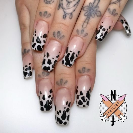 Close-up of hands with long, square-shaped nails featuring a cow-print design on a nude and white background, showcasing beauty and nail art.