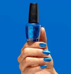 Hand holding a blue nail polish bottle against a solid blue background, showcasing matching metallic blue nail lacquer.