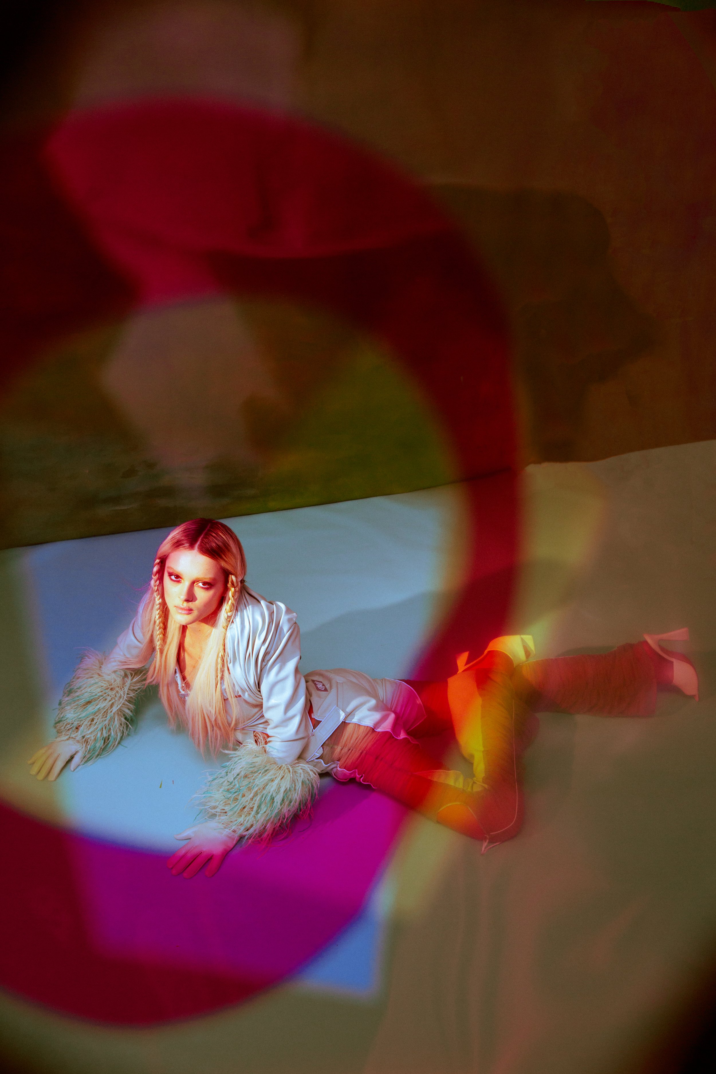Blonde female model with braids and silver top poses on a light blue surface with abstract lighting for a fashion editorial.