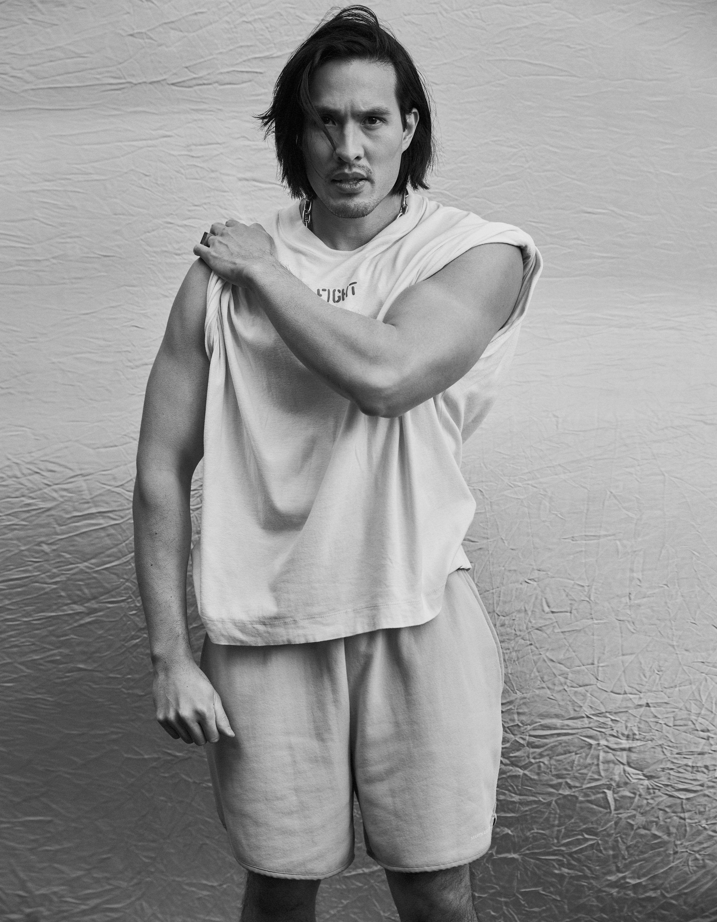 Black and white studio portrait of a muscular adult male with shoulder-length dark hair, wearing a sleeveless hoodie and shorts.