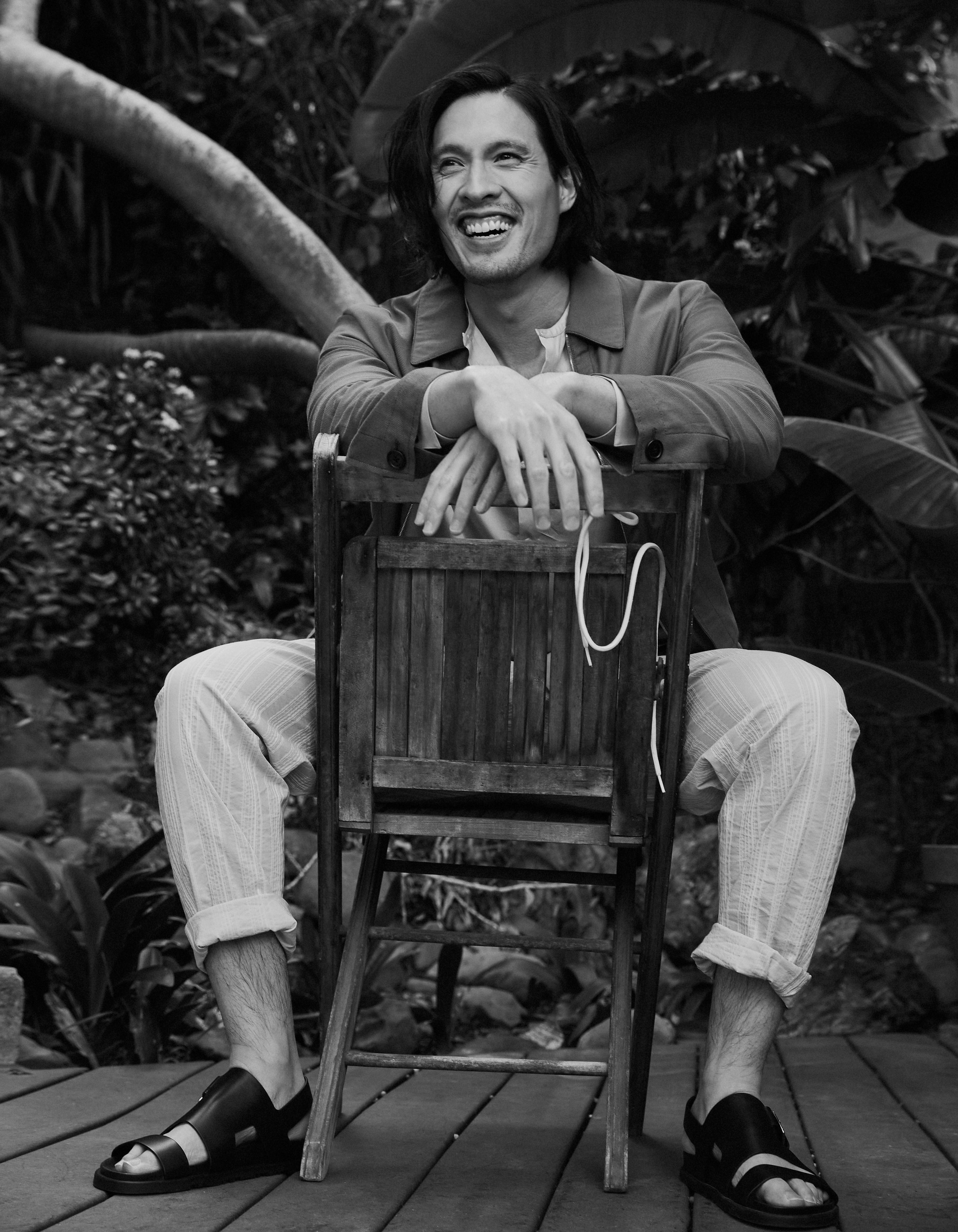 Black and white shot of a man sitting backwards on a chair outdoors, smiling, wearing a jacket, striped pants and sandals.