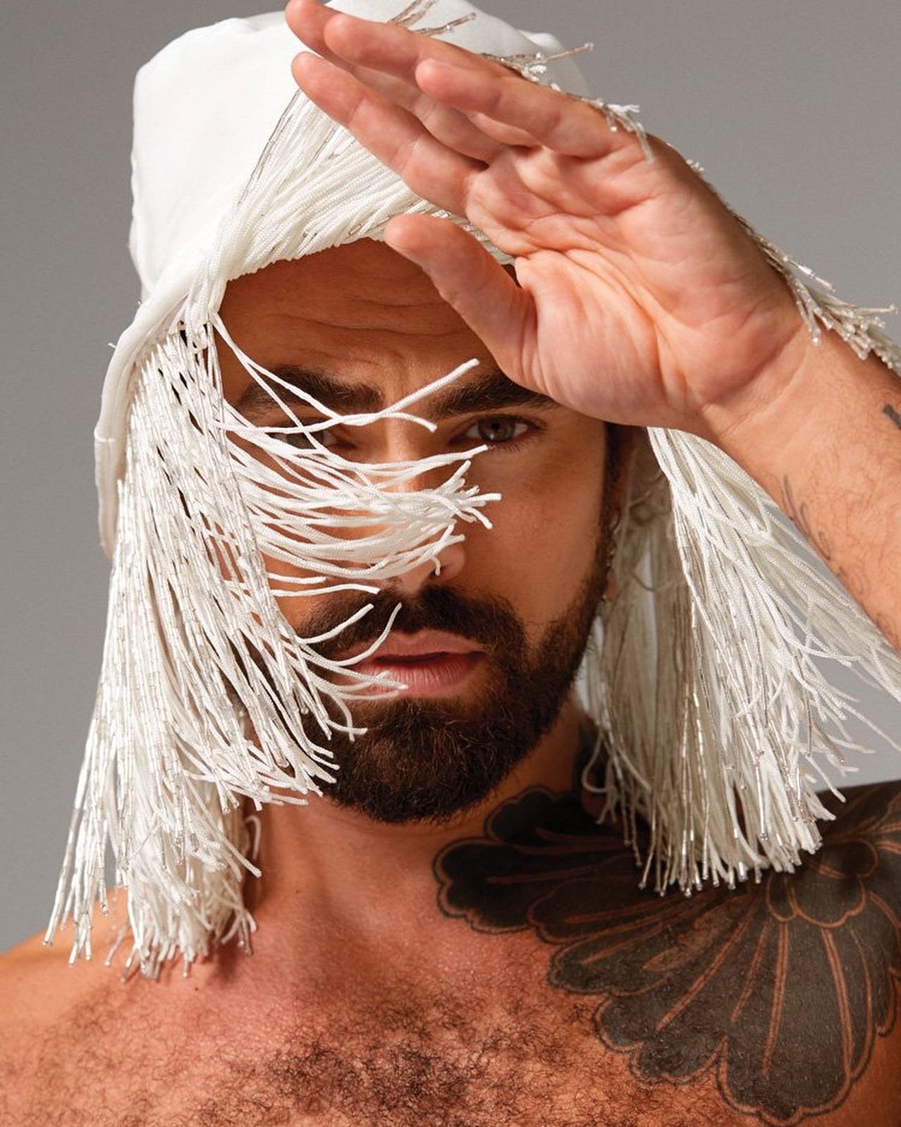 Man with beard and chest tattoo wearing white fringed headwear, covering part of his face with hand, posing against plain grey backdrop.