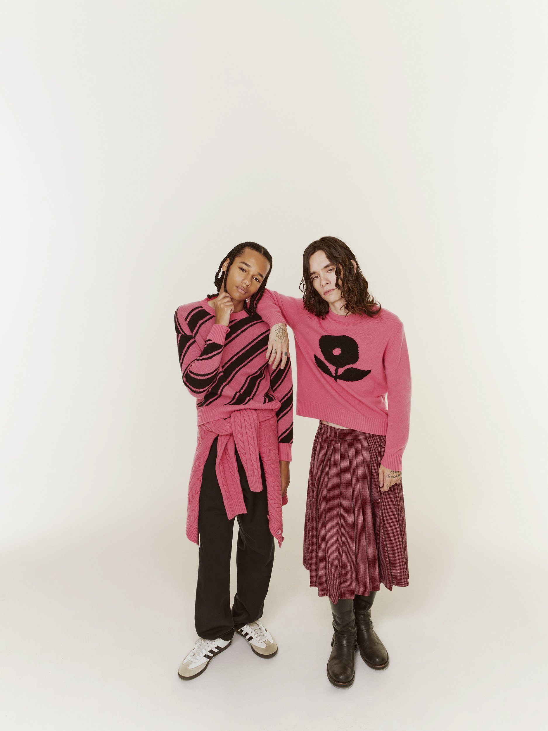 Two young adults pose in a photo studio wearing pink sweaters and casual bottoms, standing against a bright white seamless backdrop.