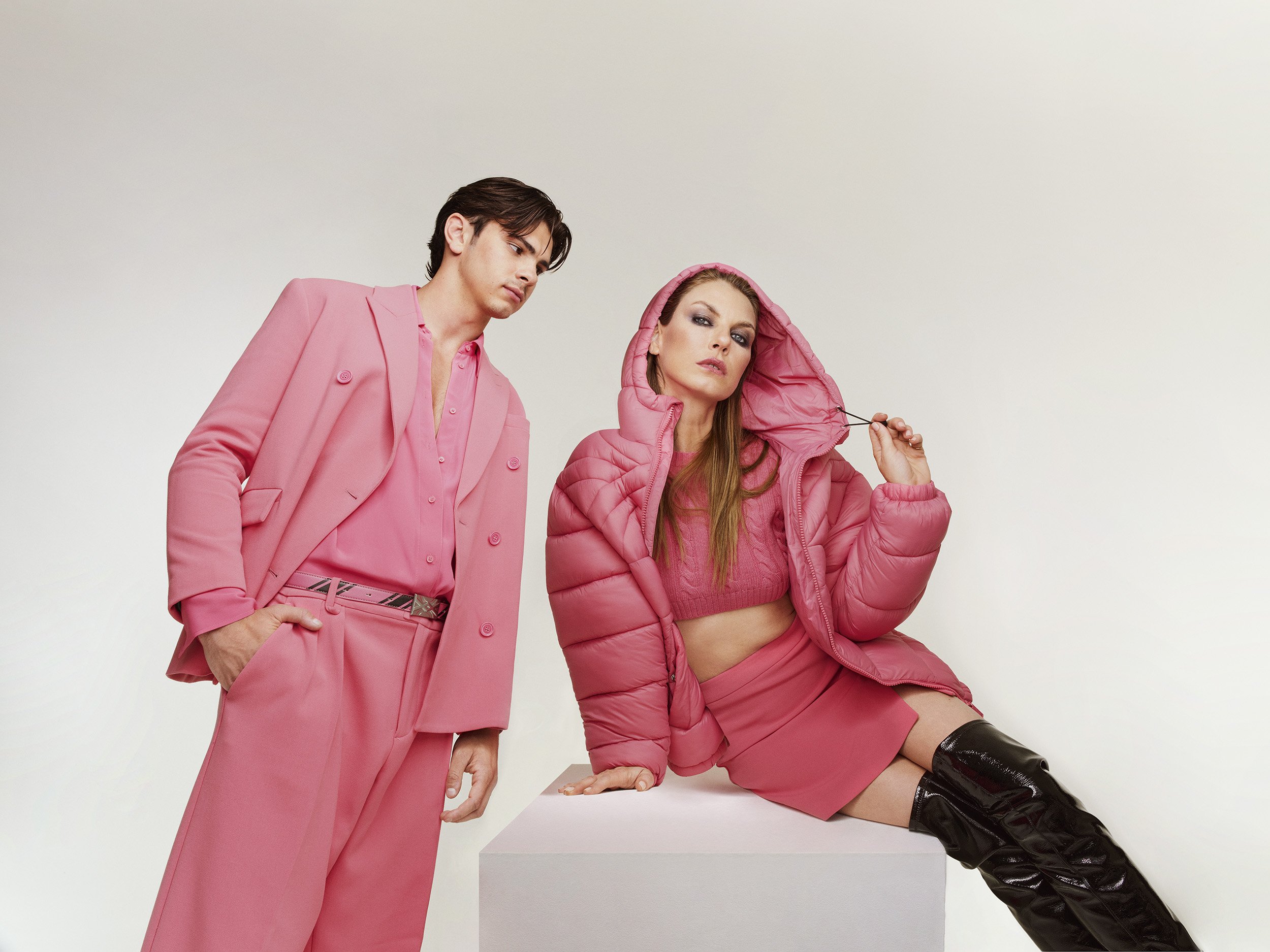 Two fashion models in pink outfits pose in a studio setting. The woman wears a puffer jacket and boots, while the man wears a suit.