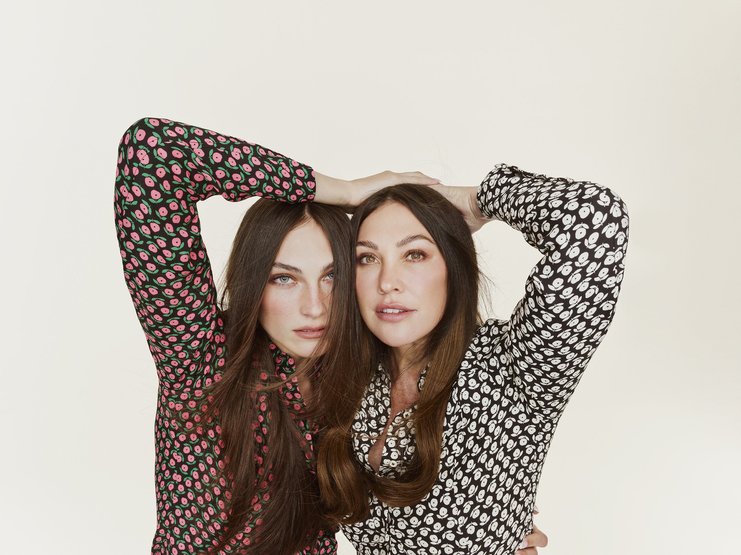 Two women pose with arms above their heads, wearing patterned long-sleeve tops against a neutral backdrop in a studio setting.