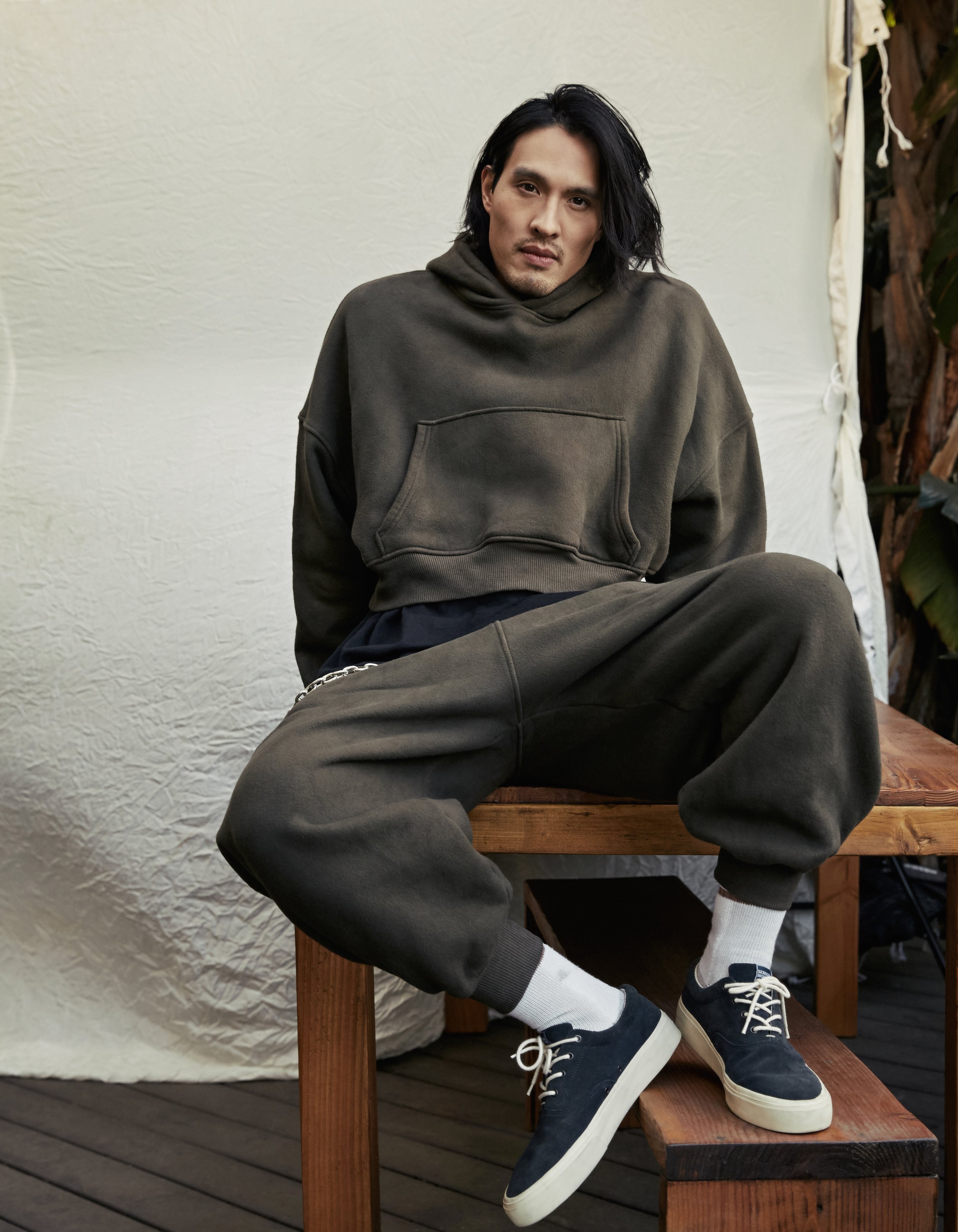Adult man wearing a dark green hoodie and sweatpants is sitting on a wood bench, looking at the camera in an indoor setting.