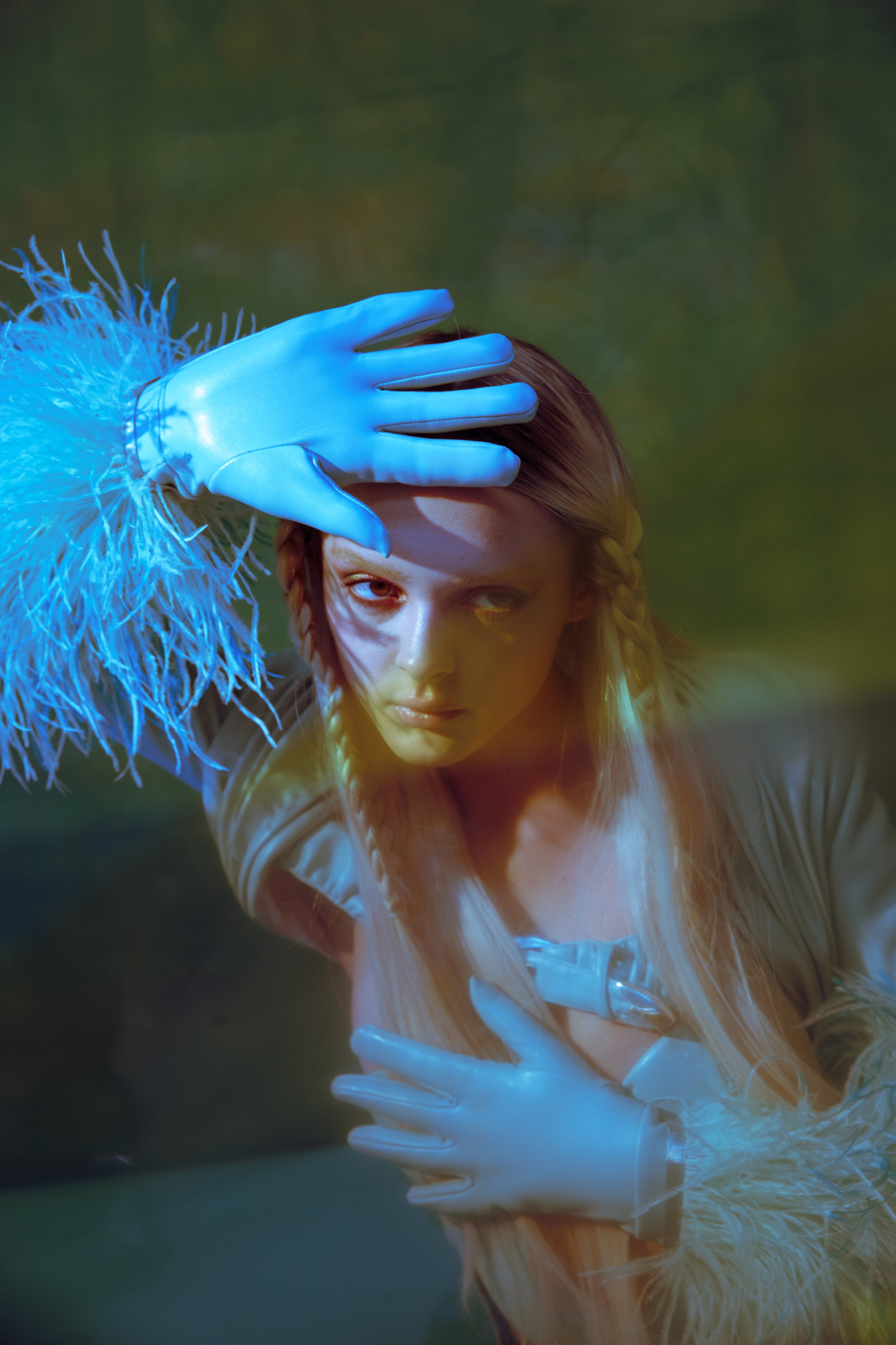 Woman in latex gloves poses in surreal, artistic portrait with feathered trim, dramatic lighting, and muted earth-tone background.