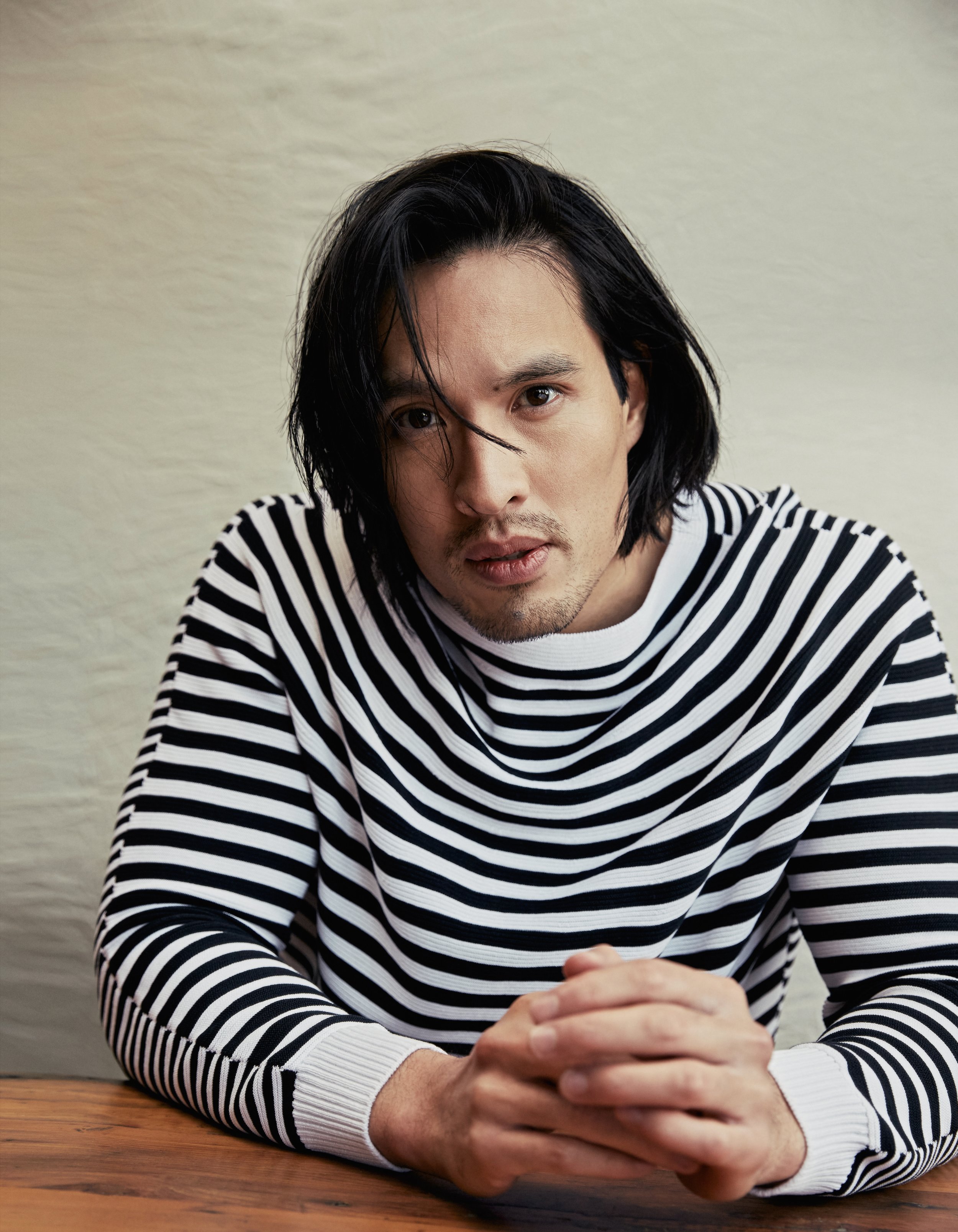 Portrait of an adult male with long dark hair, wearing a striped sweater, sitting at a wooden table, with his hands clasped in front of him.