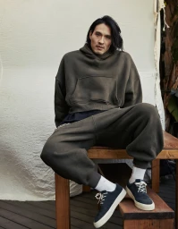 Man with long black hair wearing a dark green hoodie and matching sweatpants sitting on a wooden bench.