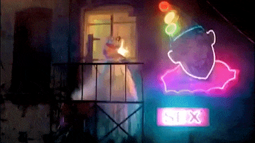 Ghostly figure on a balcony next to a neon sign of a clown with a party hat in a dimly lit, eerie indoor setting.