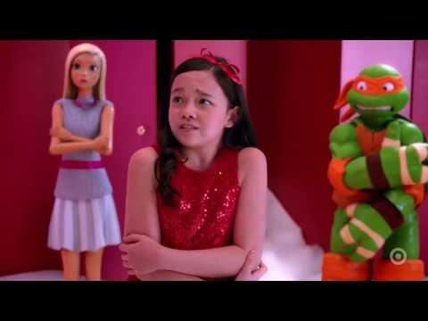 Young girl in red dress with Barbie doll and Ninja Turtle figure in a red and white room with white flooring. Bright lighting.