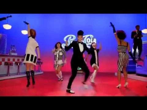 Energetic dance scene features several dancers in retro attire performing in a vibrant 1950s-style diner setting under bright lighting.