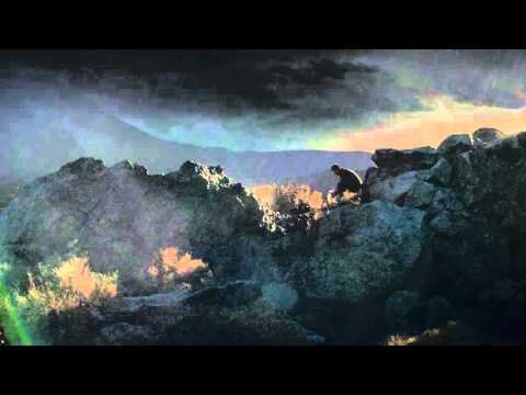 Silhouette of a man crouched on rocky terrain at sunset, overlooking a mountain range beneath a dramatic, cloudy sky. 