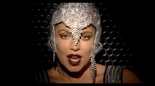 Close-up of a woman with a jeweled headpiece against a black background with honeycomb patterns. She wears sparkling jewelry.
