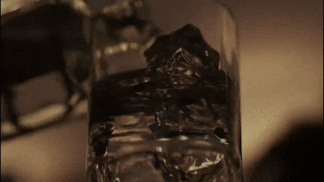 Close up view of a glass tumbler filled with ice cubes and amber liquid against a dark moody background.