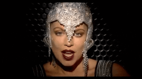 Close-up portrait of a woman wearing an ornate silver crystal-embellished headdress and dark lipstick against a dark honeycomb background.