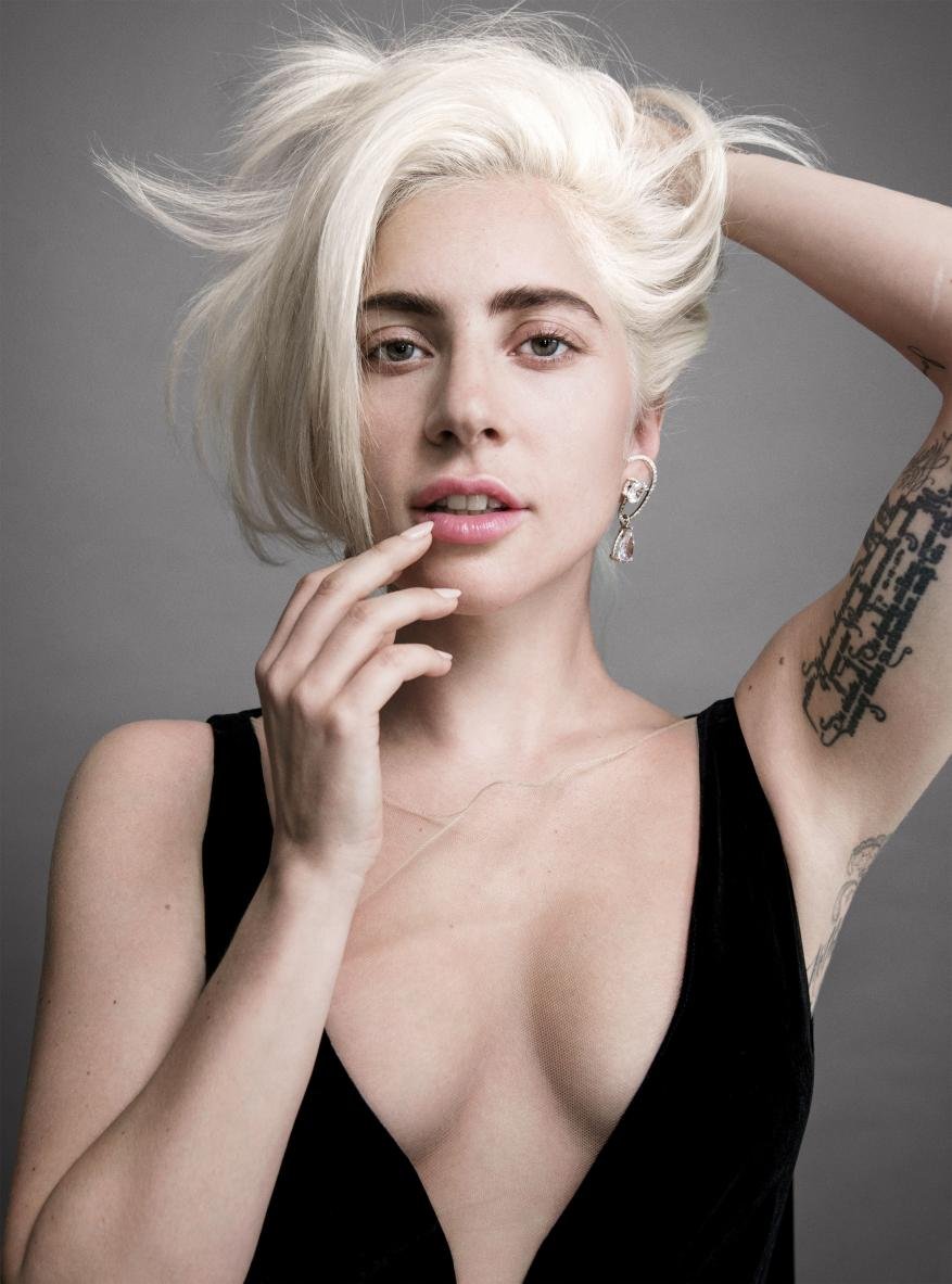 Close-up studio portrait of a fair-skinned adult female with platinum blonde hair, black V-neck dress, and geometric tattoo.